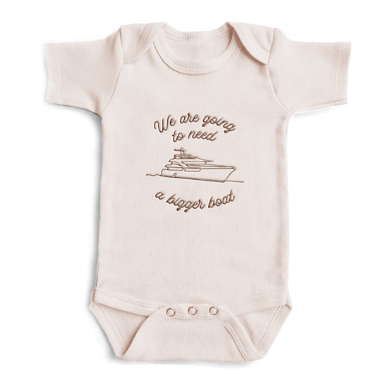 A pink colored baby bodysuit with the words "We are going to need a bigger boat" and a sea vessel embroidered on the front in gold thread