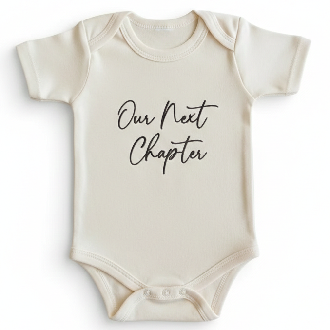 A cream colored baby bodysuit with "Our Next Chapter" embroidered on the front in black thread