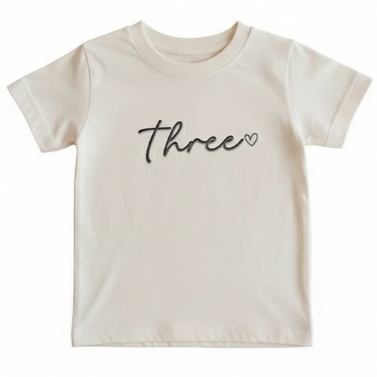 A beige T-Shirt with the word "Three" and a heart embroidered on the front in rose gold thread
