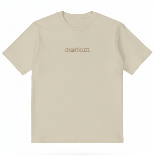 A tan T-Shirt with the word "custom" embroidered on the front in gold thread