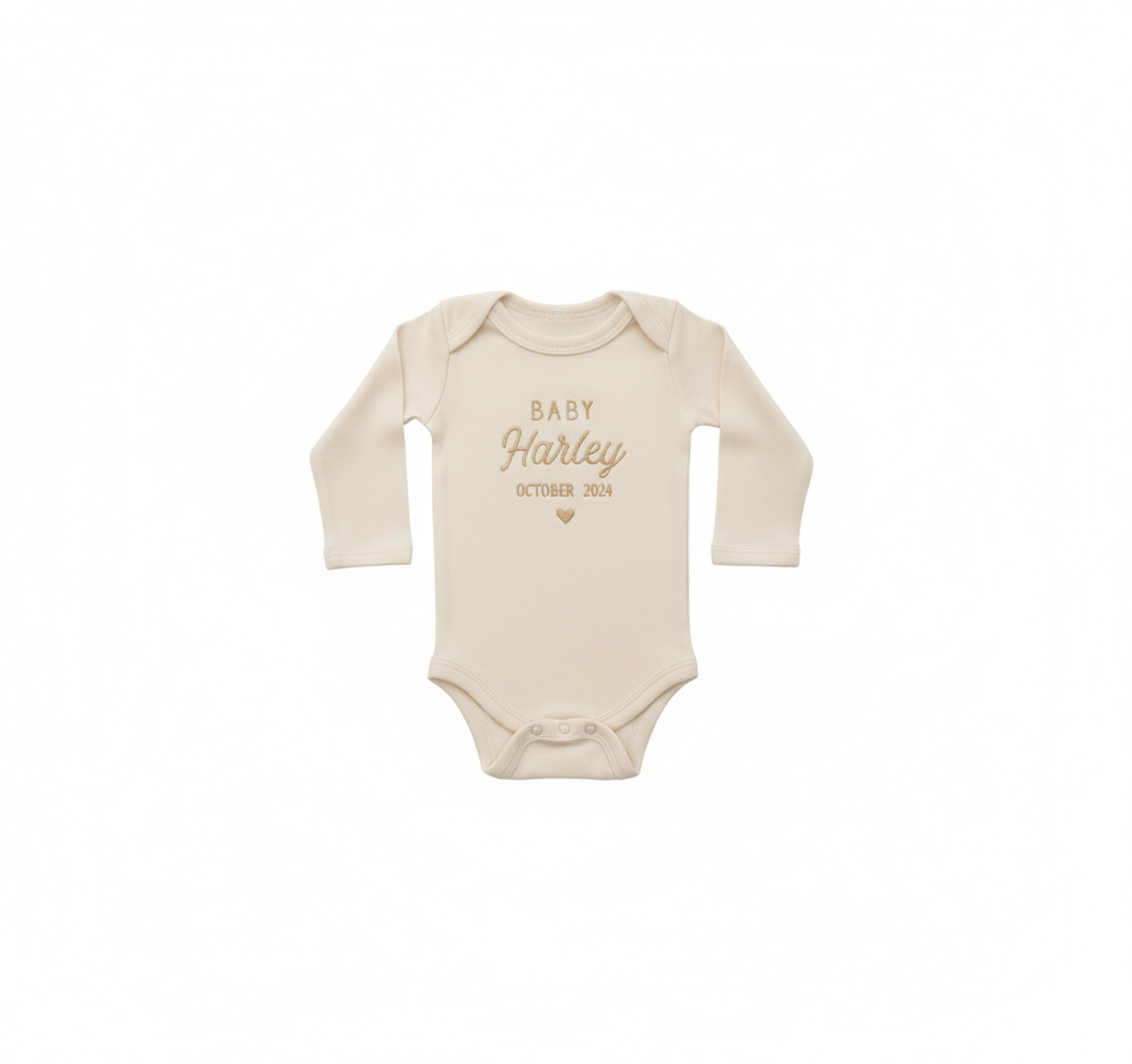 A cream colored baby bodysuit with "Baby Harley October 2024" and a heart embroidered on the front in gold thread