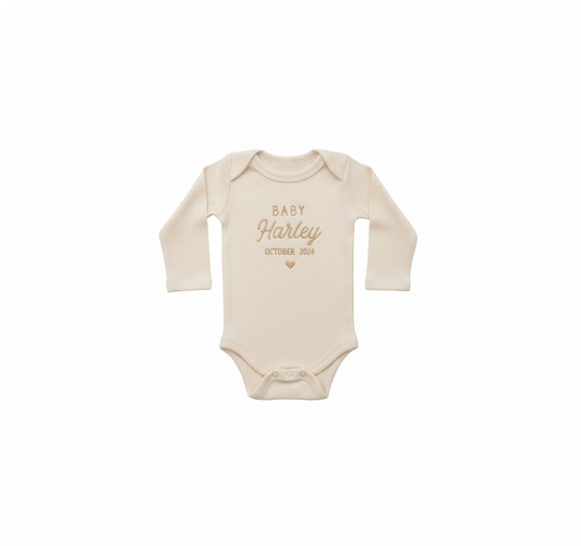 A cream colored baby bodysuit with "Baby Harley October 2024" and a heart embroidered on the front in gold thread