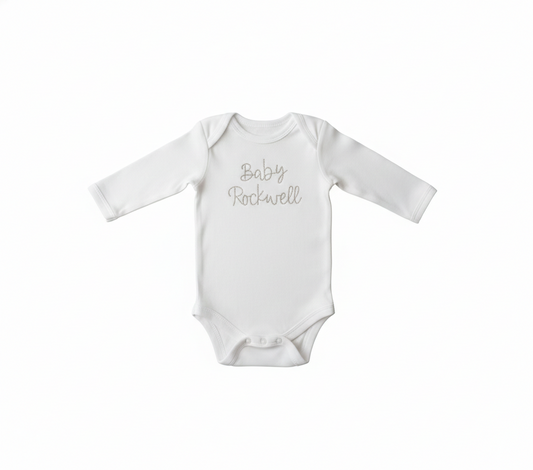 A white colored baby bodysuit with "baby rockwell" embroidered on the front in gold thread