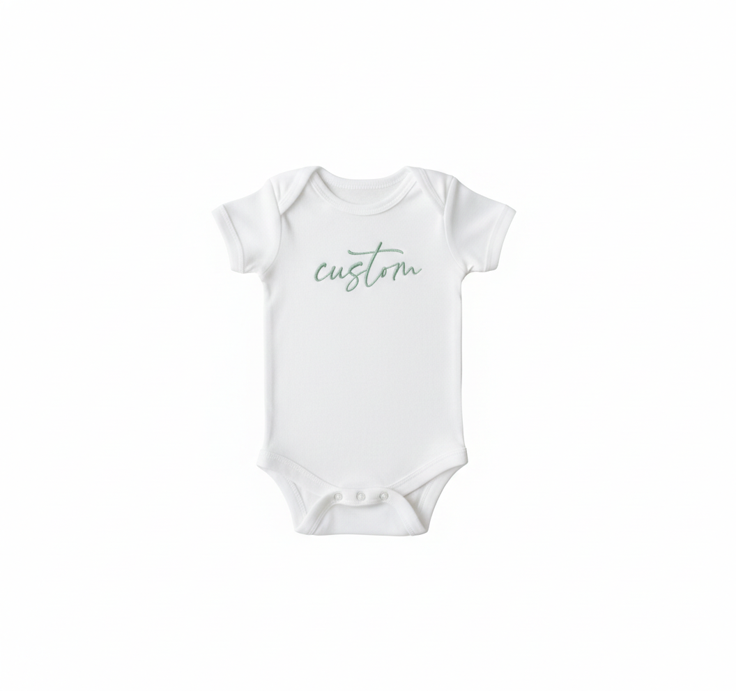 A white colored baby bodysuit with "Custom" embroidered on the front in green thread
