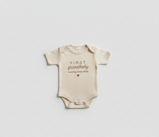 A beige colored baby bodysuit with "first grandbaby coming soon 2025" embroidered on the front in gold thread