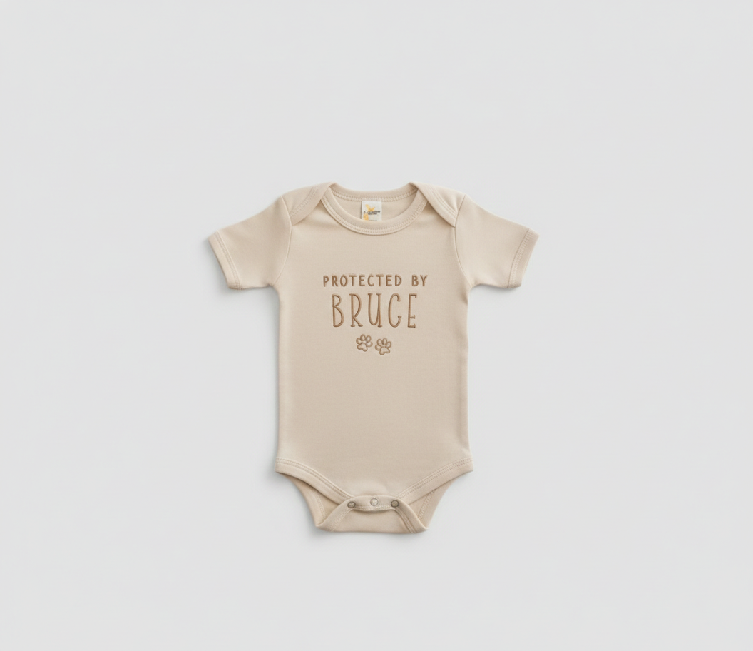 A beige colored baby bodysuit with "protected by bruce" and two paw prints embroidered on the front in gold thread

