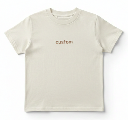 A cream colored T-Shirt with the word "custom" embroidered on the front in bronze thread