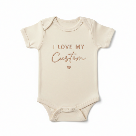 A cream colored baby bodysuit with "I Love My Custom" embroidered on the front in gold thread