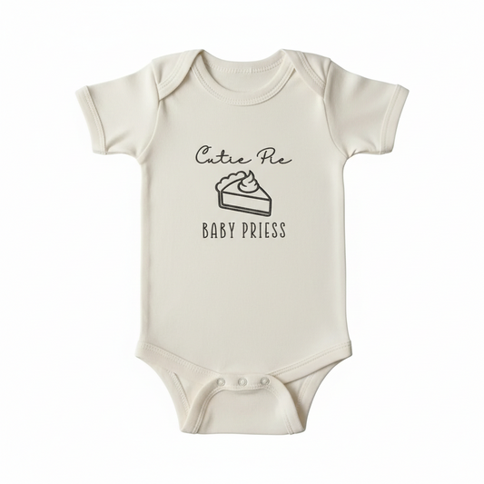 A cream colored baby bodysuit with "Cutie pie Baby Priess" and a slice of pie embroidered on the front in brown thread