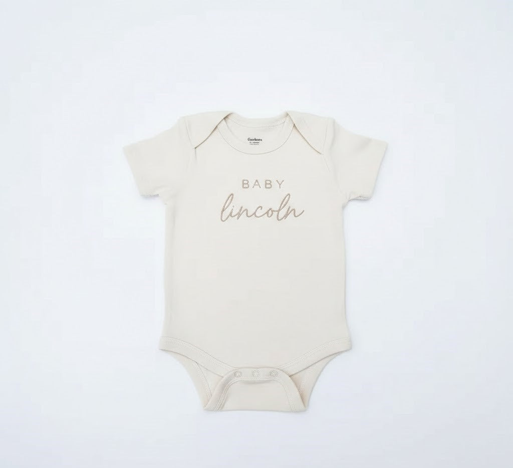 A cream baby bodysuit with the words "baby lincoln" embroidered on the front in gold thread