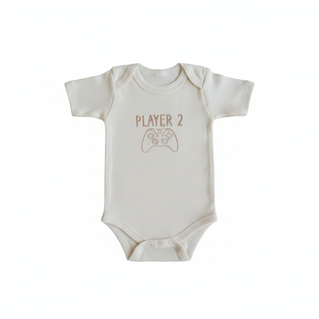 A cream colored baby bodysuit with "Player 2" and a gaming console controller embroidered on the front in gold thread