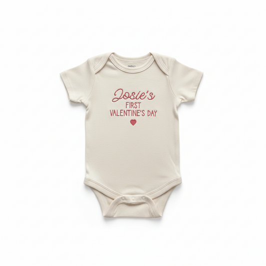 A cream colored baby bodysuit with "Josie's First Valentine's Day" embroidered on the front in pink thread