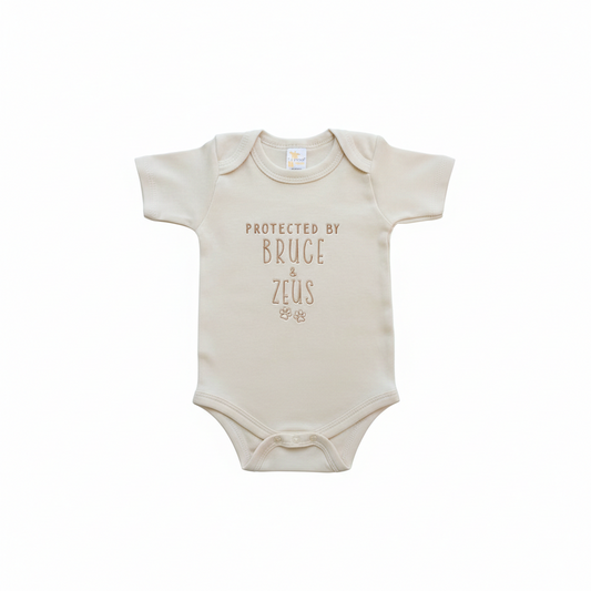 a cream colored baby bodysuit with the text "Protected by bruce and zeus" and two paw prints embroidered on the front with gold thread