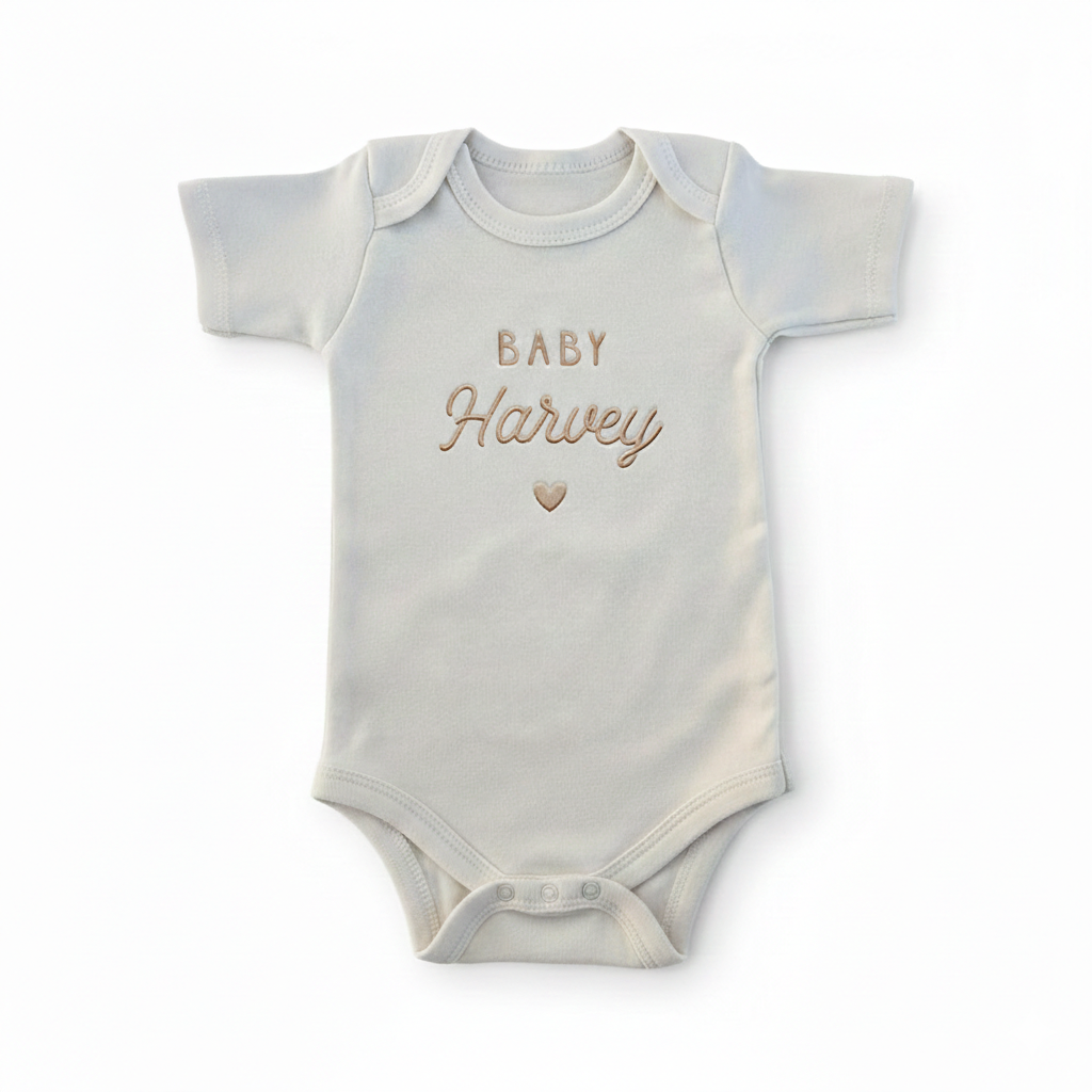 A cream colored baby bodysuit with "baby harvey" and a heart embroidered on the front in gold thread
