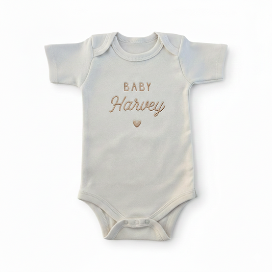 A cream colored baby bodysuit with "baby harvey" and a heart embroidered on the front in gold thread