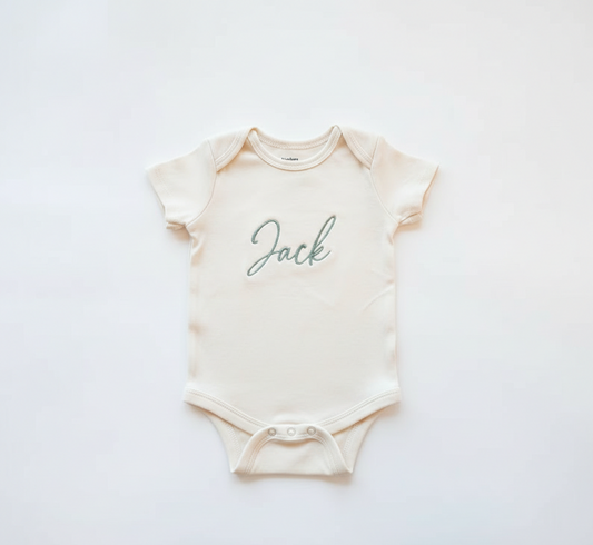 A cream colored baby onesie with the name "Jack" embroidered in green thread on the front.