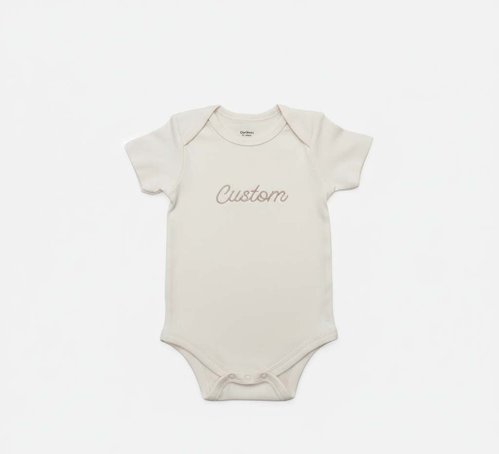 A cream colored baby bodysuit with "Custom" embroidered on the front in gold thread