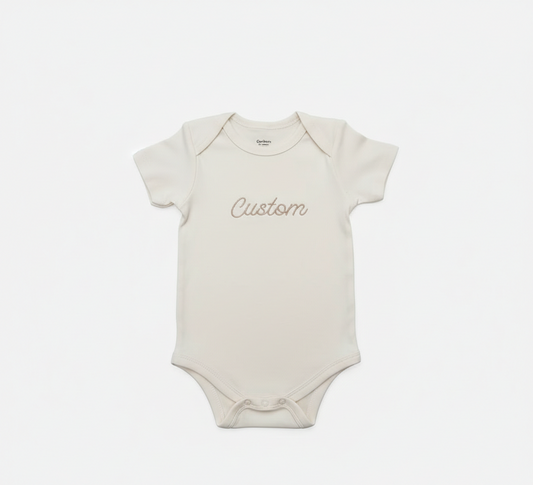 A cream colored baby bodysuit with "Custom" embroidered on the front in gold thread