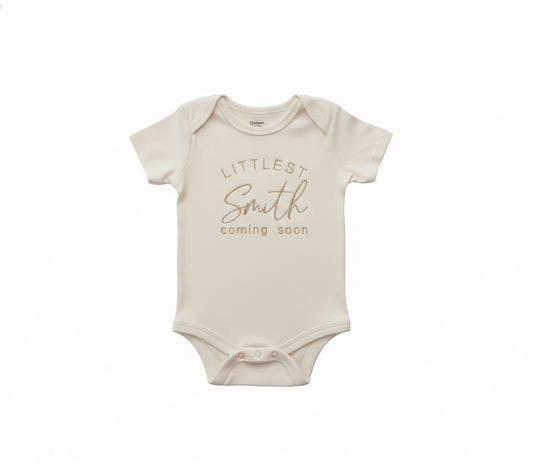 A cream colored baby bodysuit with "Littlest Smith Coming Soon" embroidered on the front in gold thread
